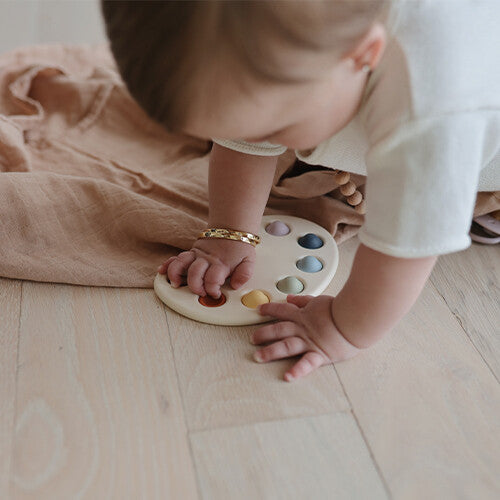Een baby kruipt en raakt het mushie sensorisch verfpalet aan op de houten vloer.