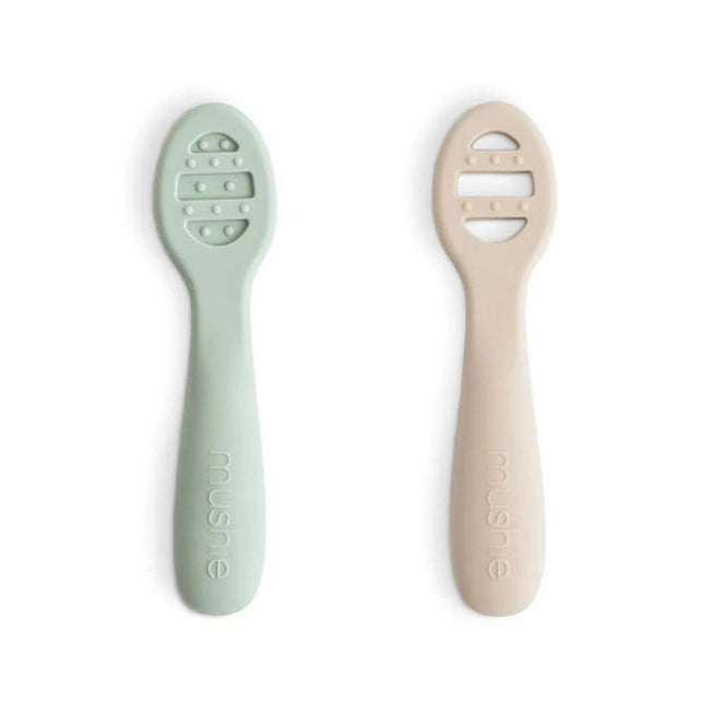 Twee mushie sensorische babylepels, een cambridge blue en een shifting sands, liggen naast elkaar op een witte achtergrond.