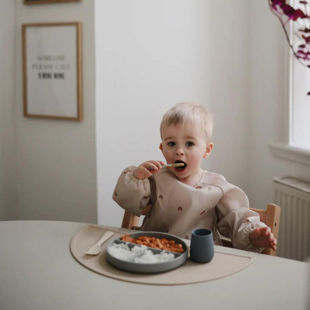 Een baby in een kinderstoel eet met een mushie sensorische babylepel van een bord met twee vakken.