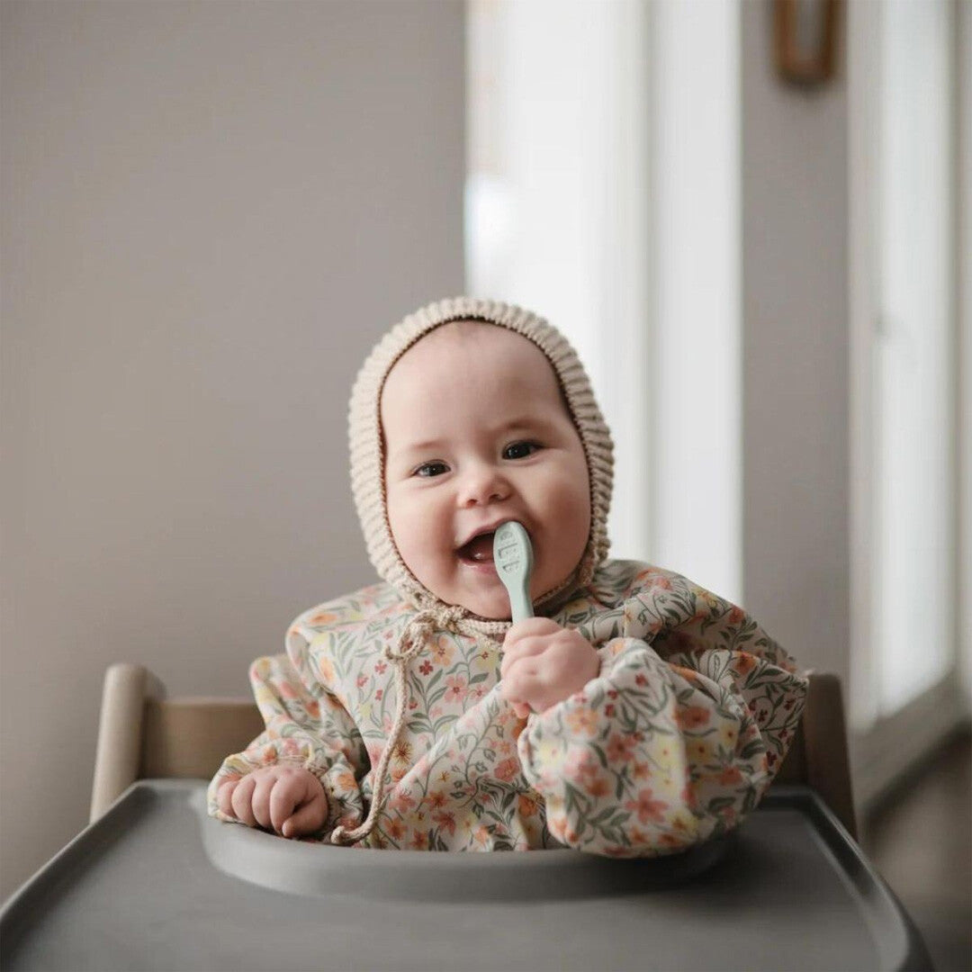 Een lachende baby met een bloemen slabber en mutsje houdt een mushie sensorische babylepel in de mond.