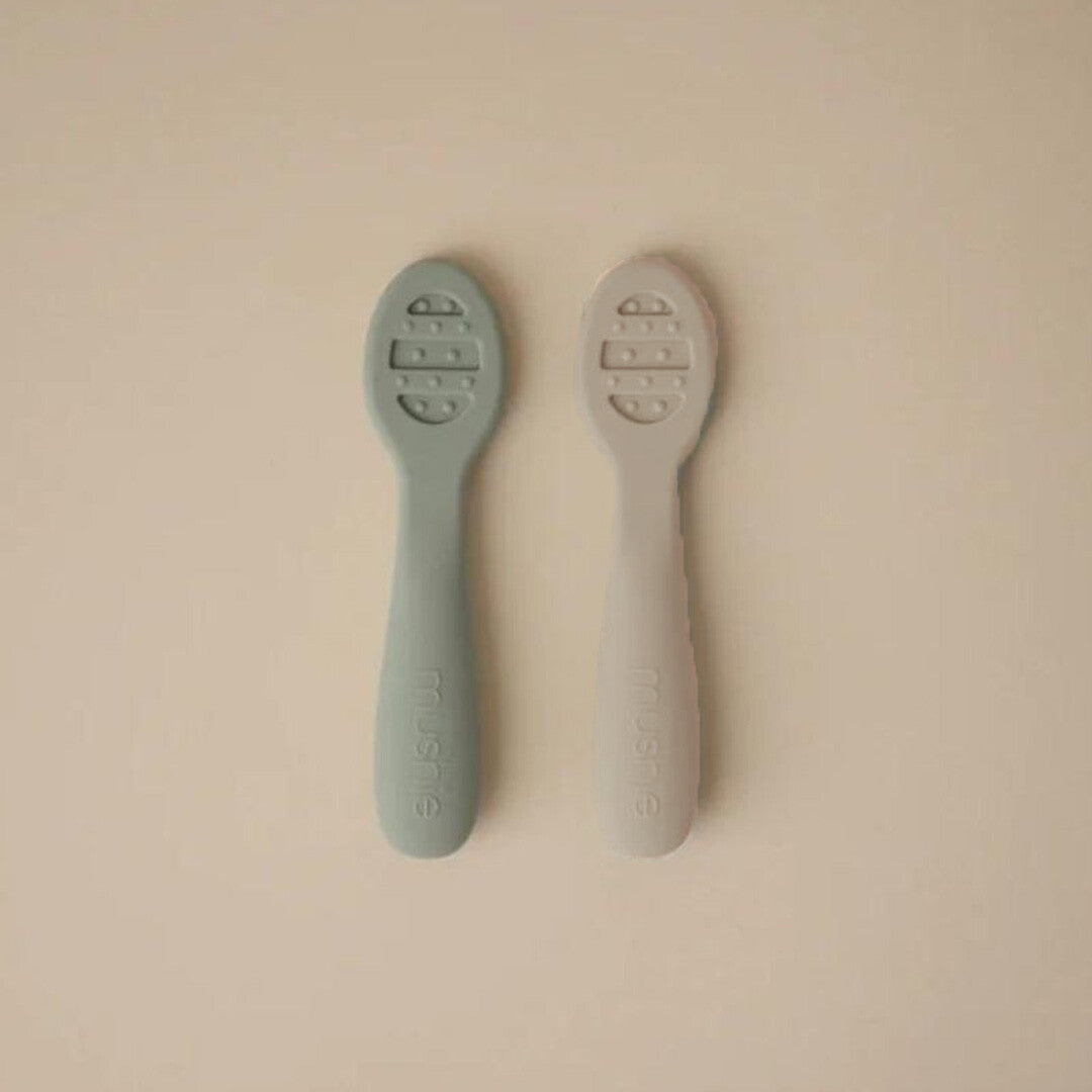Twee mushie sensorische babylepels, een cambridge blue en een shifting sands, liggen naast elkaar op een lichte achtergrond.
