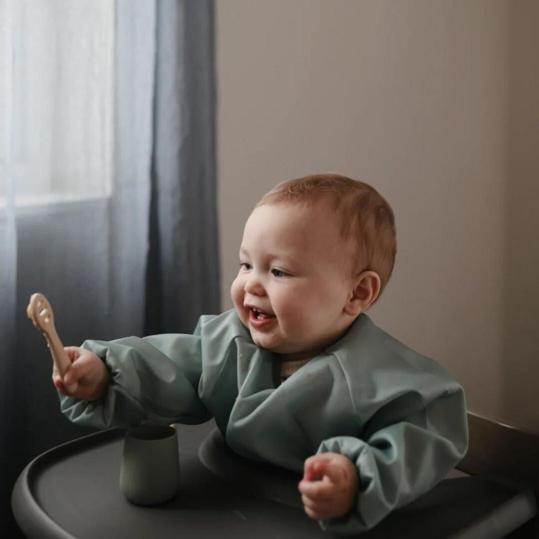 Een lachende baby in een kinderstoel die een Mushie sensorische babylepel vasthoudt.
