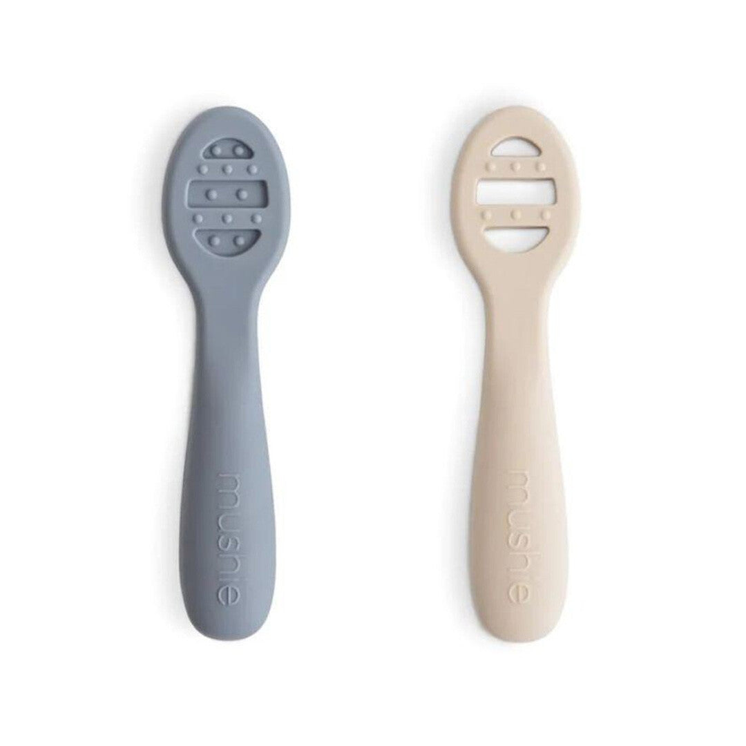 Twee Mushie siliconen sensorische babylepels in grijsblauw en beige.