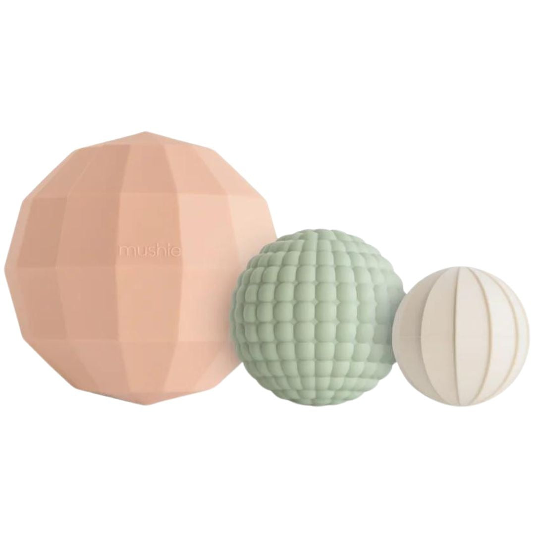 Drie mushie sensorische ballen in verschillende maten en texturen, in de kleuren perzik, lichtgroen en crème.