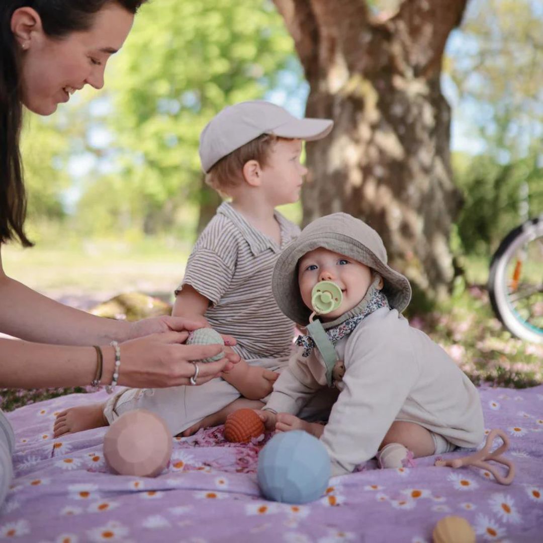 Een vrouw en twee baby's spelen buiten op een deken met mushie sensorische ballen.