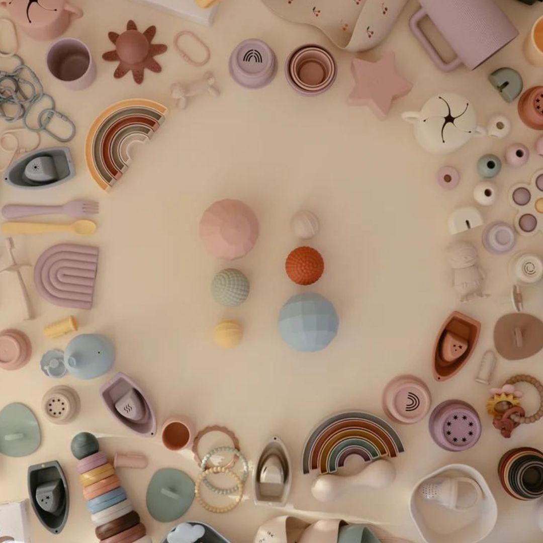 Een overzicht van diverse mushie babyproducten, waaronder sensorische ballen, stapelspeelgoed en bijtringen, in een cirkel gerangschikt.