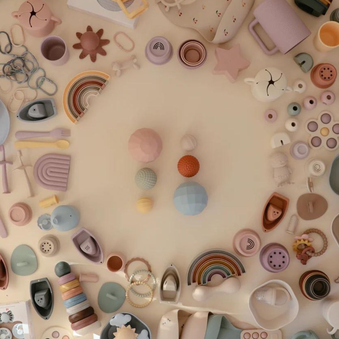 Een bovenaanzicht van een collectie mushie babyproducten, waaronder sensorische ballen, in verschillende kleuren en vormen.