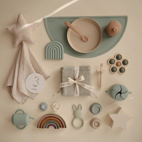 Een flat lay van diverse mushie babyproducten, waaronder een sensorische bloem, servies en een regenboog stapelaar.