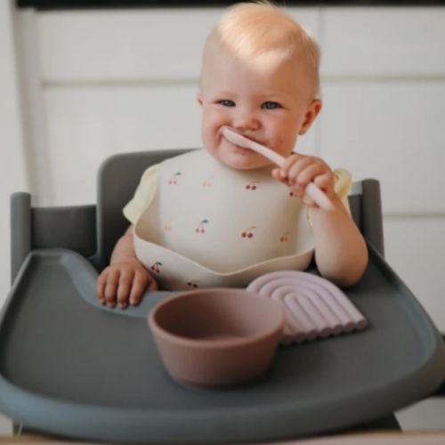 Een lachende baby in een kinderstoel houdt een Mushie siliconen babylepel bij de mond.