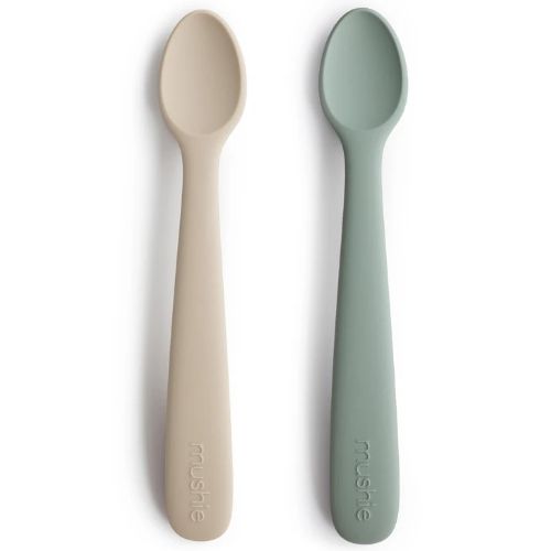 Twee Mushie siliconen babylepels in beige en lichtgroen liggen naast elkaar op een witte ondergrond.