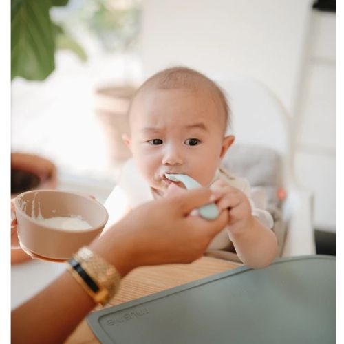 Een baby in een kinderstoel wordt gevoed met een Mushie siliconen babylepel uit een beige kom.