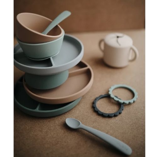 Een Mushie collectie van siliconen babyvoedingsartikelen, waaronder borden, kommen en een lepel in beige en groen.
