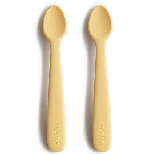 Twee mushie siliconen babylepels in de kleur pale daffodil liggen naast elkaar op een witte ondergrond.