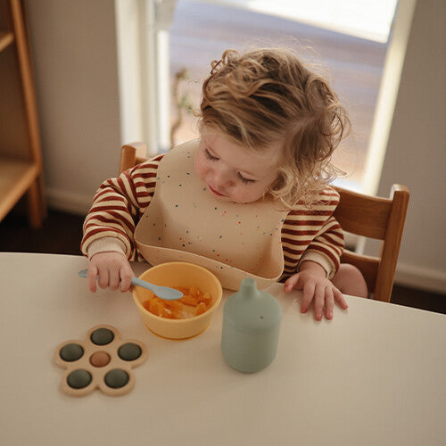 Een peuter met een slabber zit aan tafel en kijkt naar een gele kom met eten en een lichtblauwe lepel.