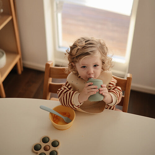 Een peuter drinkt uit een groene Mushie drinkbeker tijdens het eten, met een babylepel en kom op tafel.