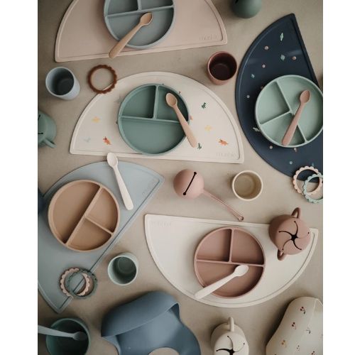 Een bovenaanzicht van een collectie Mushie siliconen babyvoedingsartikelen, waaronder placemats, borden, bekers en lepels in pastelkleuren.