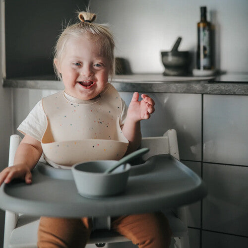 Een lachende baby zit in een kinderstoel met een slabbetje, kom en lepel.