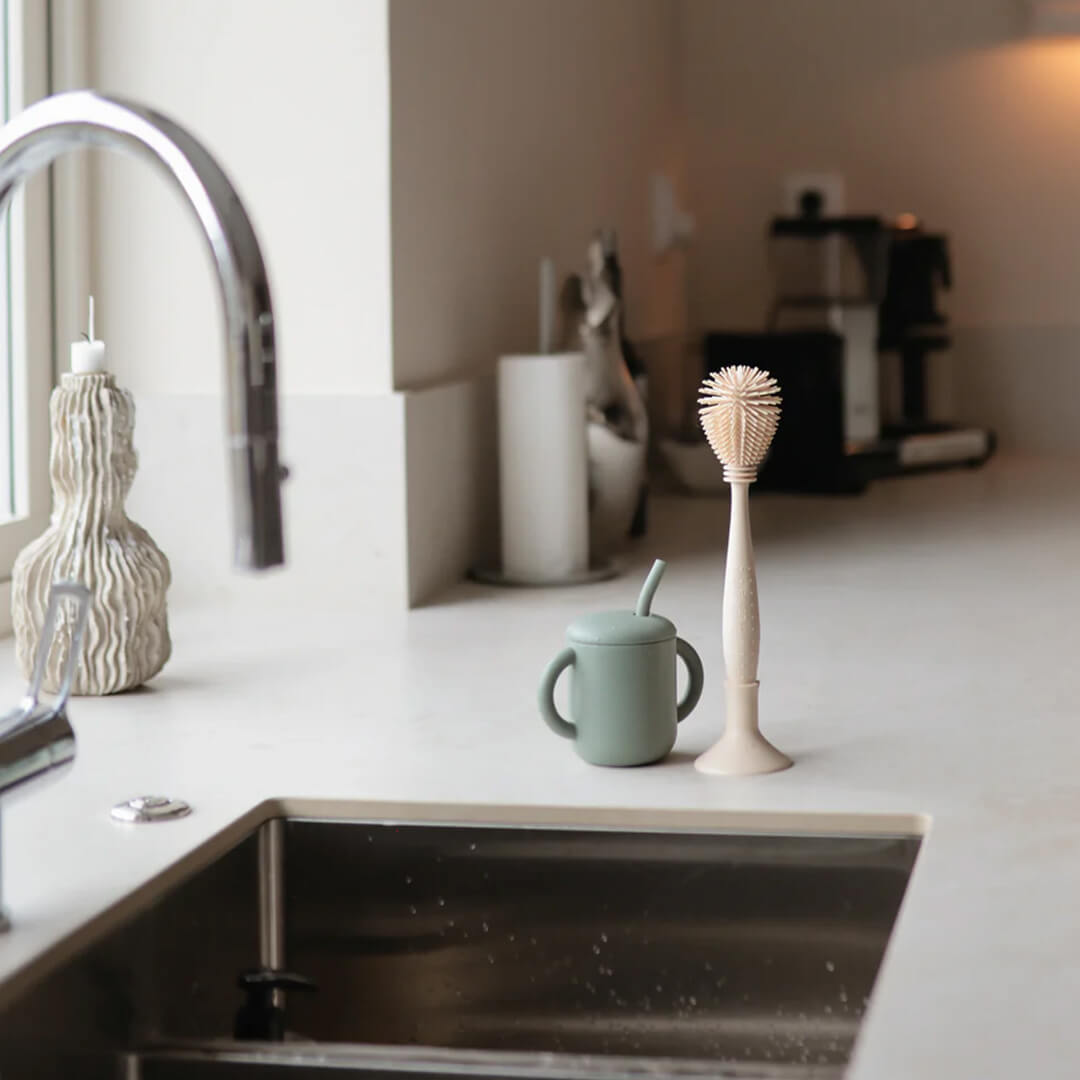 Een mushie siliconen flessenborstel in blush kleur staat naast een groene drinkbeker op een aanrecht.