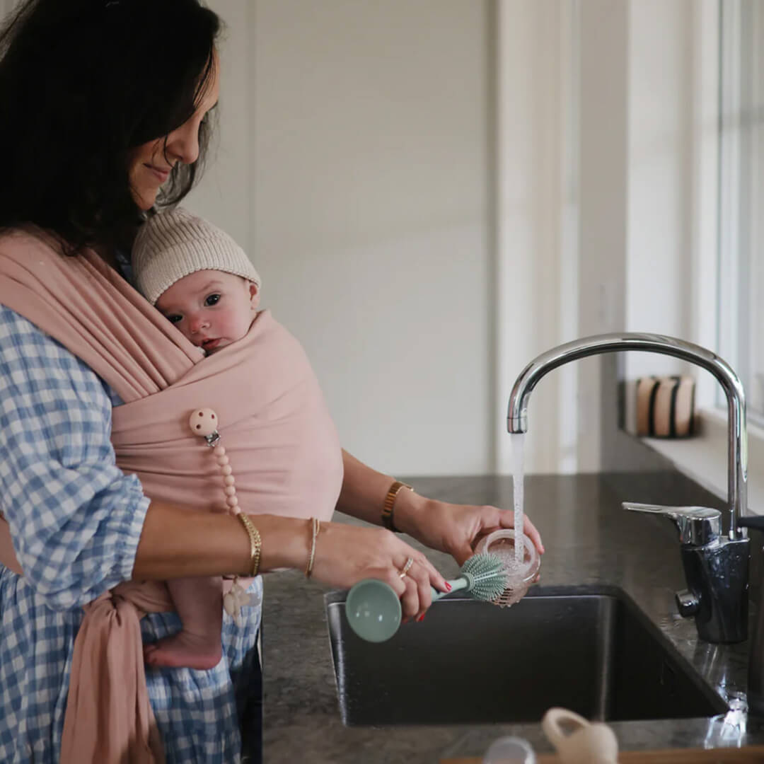 Een vrouw met een baby in een draagdoek wast een babyfles met een groene flessenborstel in de gootsteen.