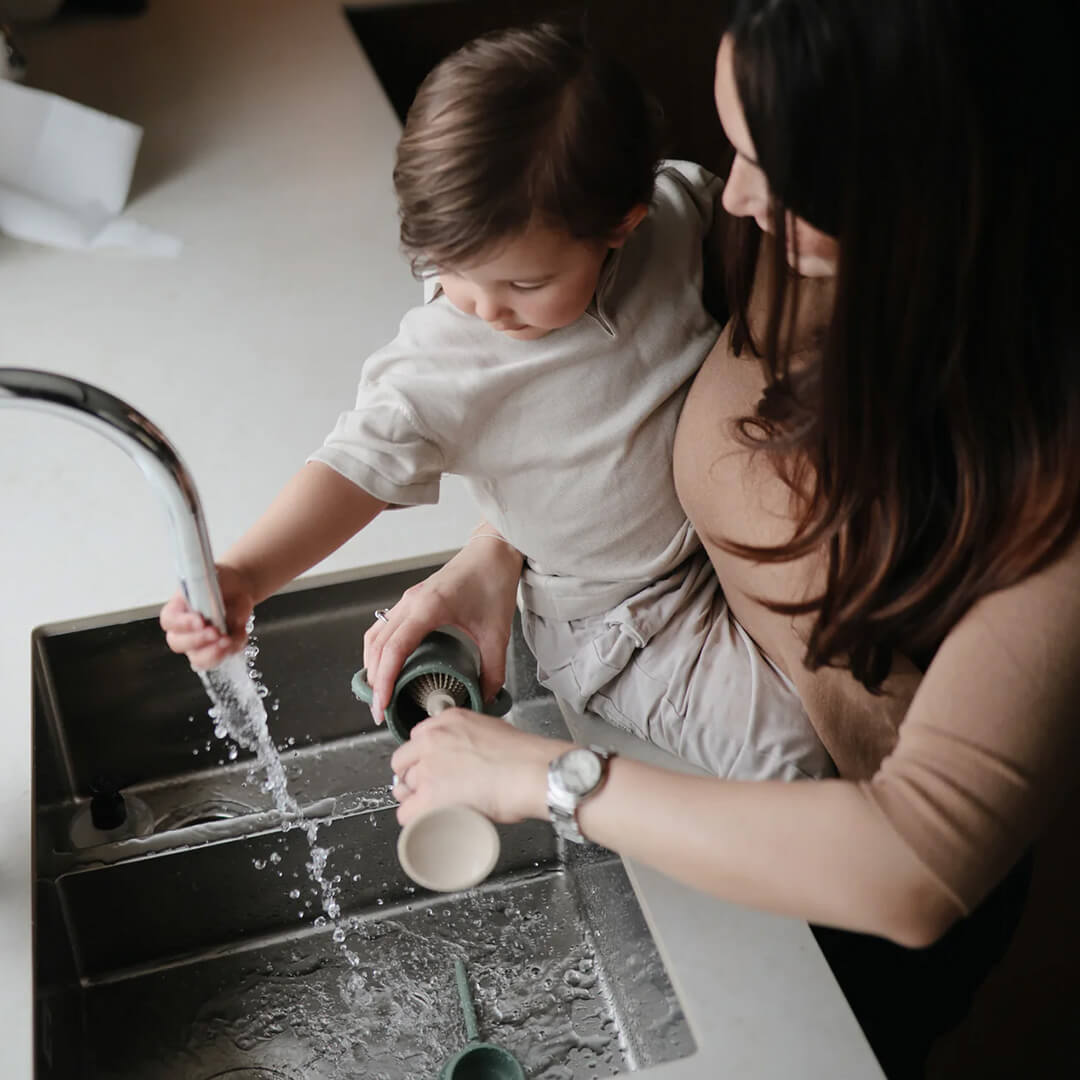 Een vrouw met een peuter op haar arm wast een babybeker met een groene flessenborstel onder de kraan.