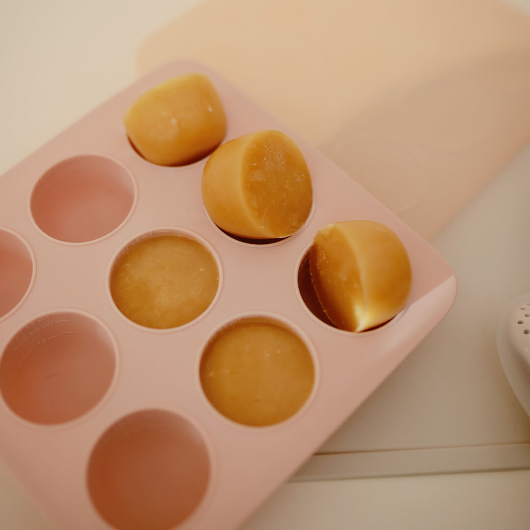 Een mushie roze siliconen invriesvorm voor babyvoeding met enkele bevroren blokjes babyvoeding.