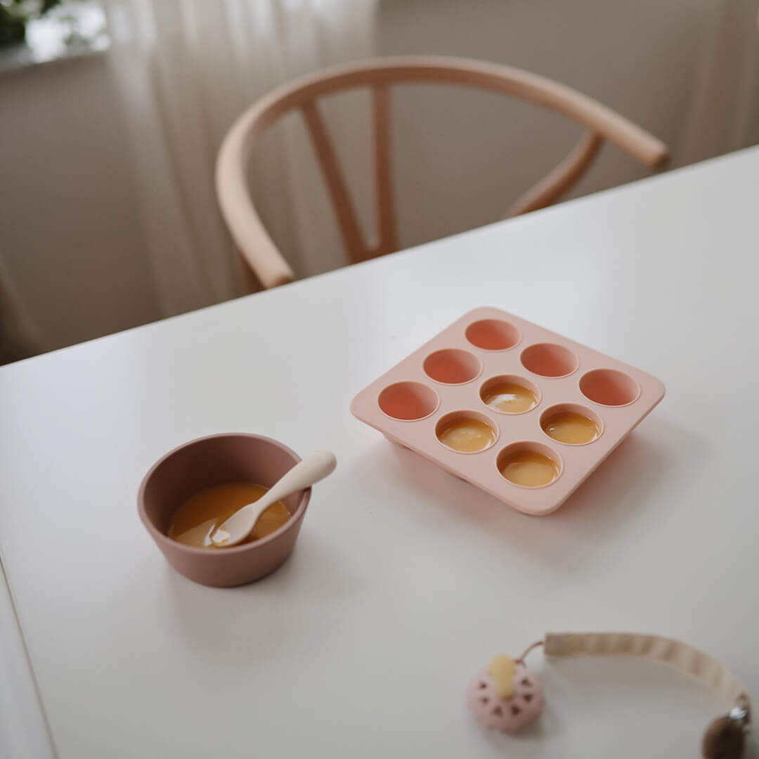 Een mushie roze siliconen invriesvorm voor babyvoeding gevuld met puree, naast een kommetje en lepel op een witte tafel.