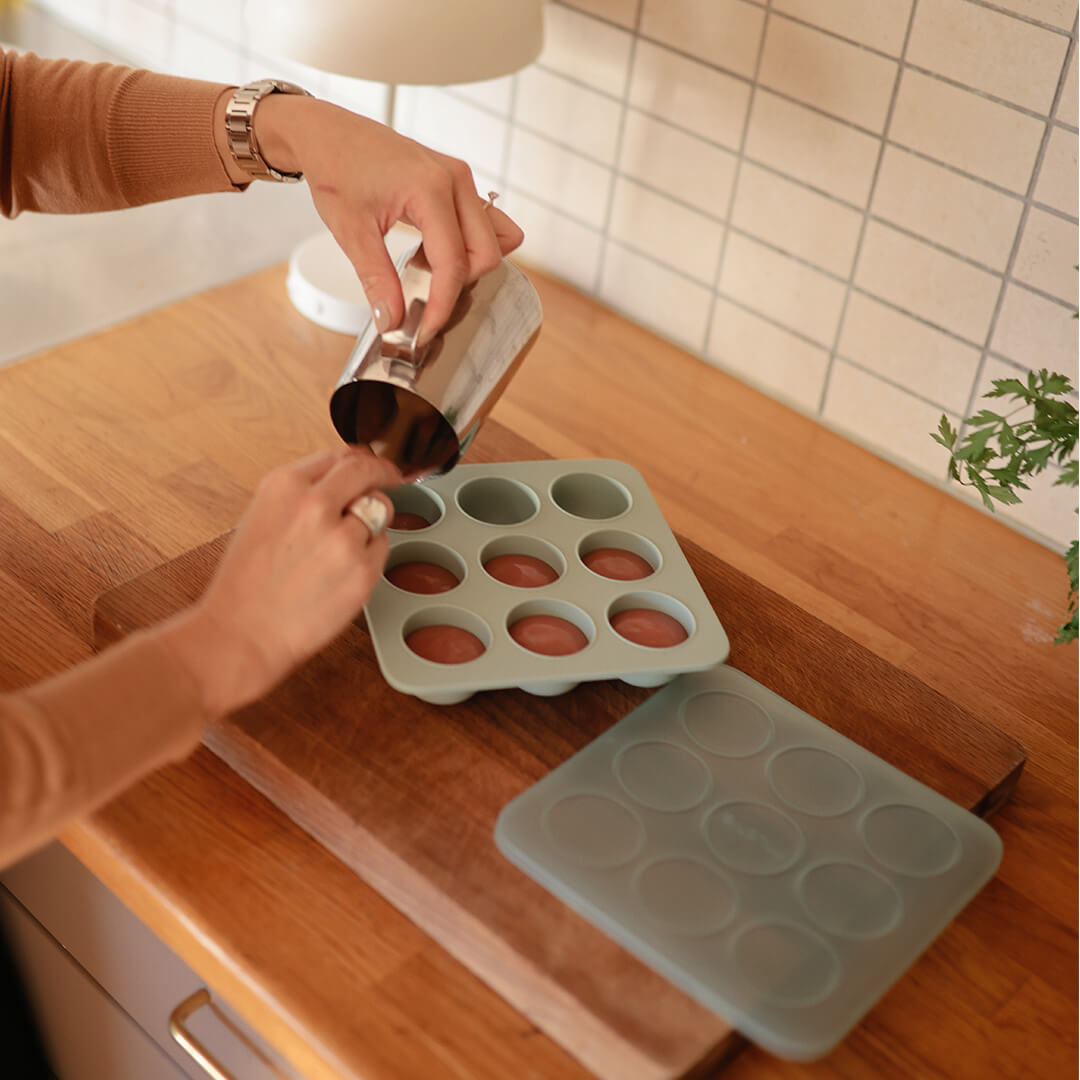 Handen vullen een Cambridge Blue mushie siliconen invriesvorm voor babyvoeding met vloeistof op een houten aanrecht.