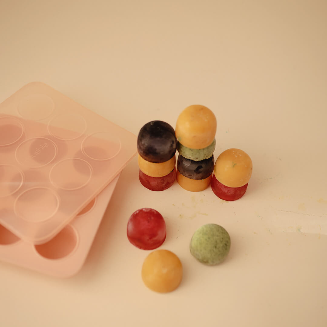 Meerdere kleurrijke, ronde bevroren babyvoedingblokjes naast een roze mushie siliconen invriesvorm en deksel.
