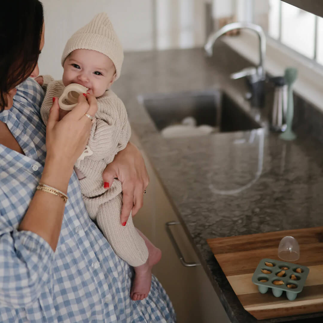 Een baby wordt gevoed met een fruitspeen, met op de voorgrond een mushie invriesvorm met voedingsblokjes.