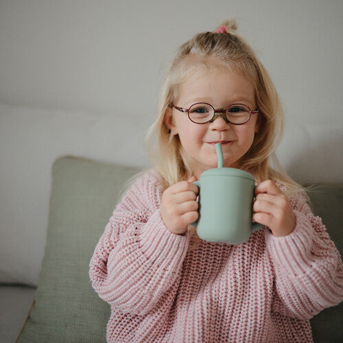 Een lachend meisje met bril drinkt uit een groene mushie siliconen kinderbeker met rietje.