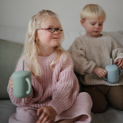 Een meisje en een jongetje zitten samen en houden elk een mushie siliconen kinderbeker met rietje vast.