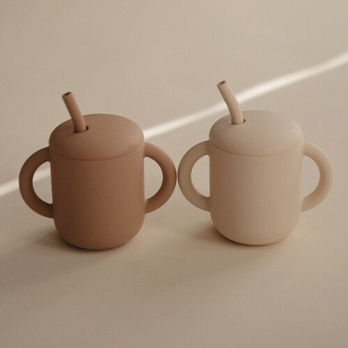Twee mushie siliconen kinderbekers met rietje in beige en lichtbeige op een lichte ondergrond.