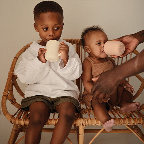 Twee kinderen drinken uit een mushie siliconen kinderbeker met rietje, zittend op een rieten stoel.
