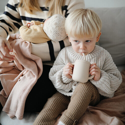 Een jongen drinkt uit een mushie siliconen kinderbeker met rietje, zittend naast een volwassene met een baby.