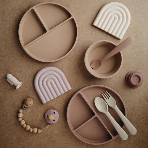 Collectie Mushie babyvoedingsproducten en accessoires, waaronder borden, een kom, bestek en bijtspeelgoed in aardetinten.