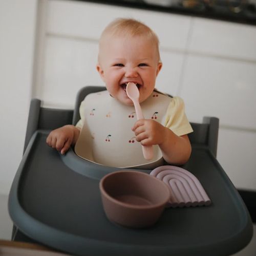 Een lachende baby in een kinderstoel met een roze lepel en een kommetje op het dienblad.