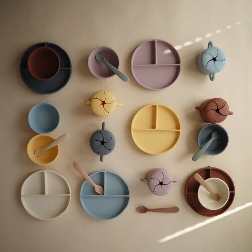 Een overzicht van diverse mushie babyvoedingsartikelen zoals borden, kommen, lepels en snackbekers in verschillende pastelkleuren.
