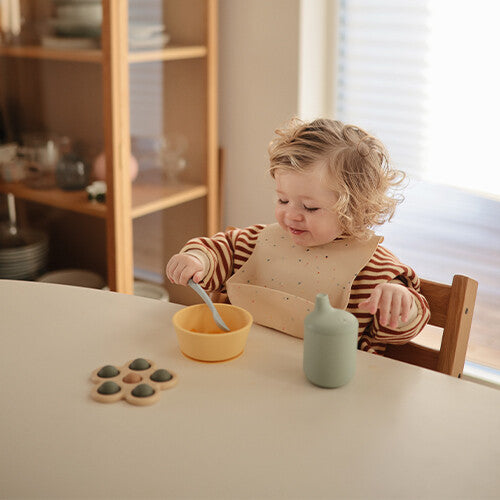 Een peuter met een slabber eet aan tafel uit een gele kinderkom.