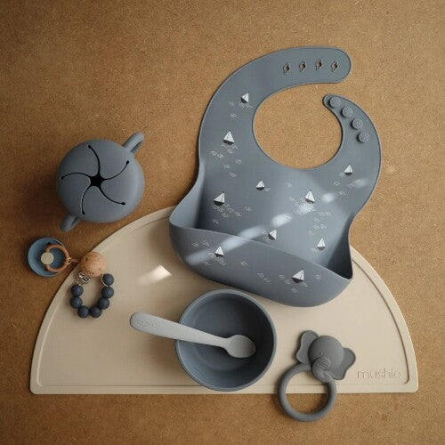 Een mushie babyvoedingsset met een grijze slab met bootjespatroon, een kom, lepel, snackcup en bijtring op een beige placemat.