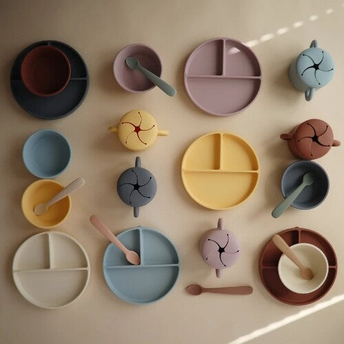Een overzicht van diverse mushie siliconen babyvoedingsbenodigdheden in verschillende pastelkleuren, waaronder kommen, borden en snackcups.