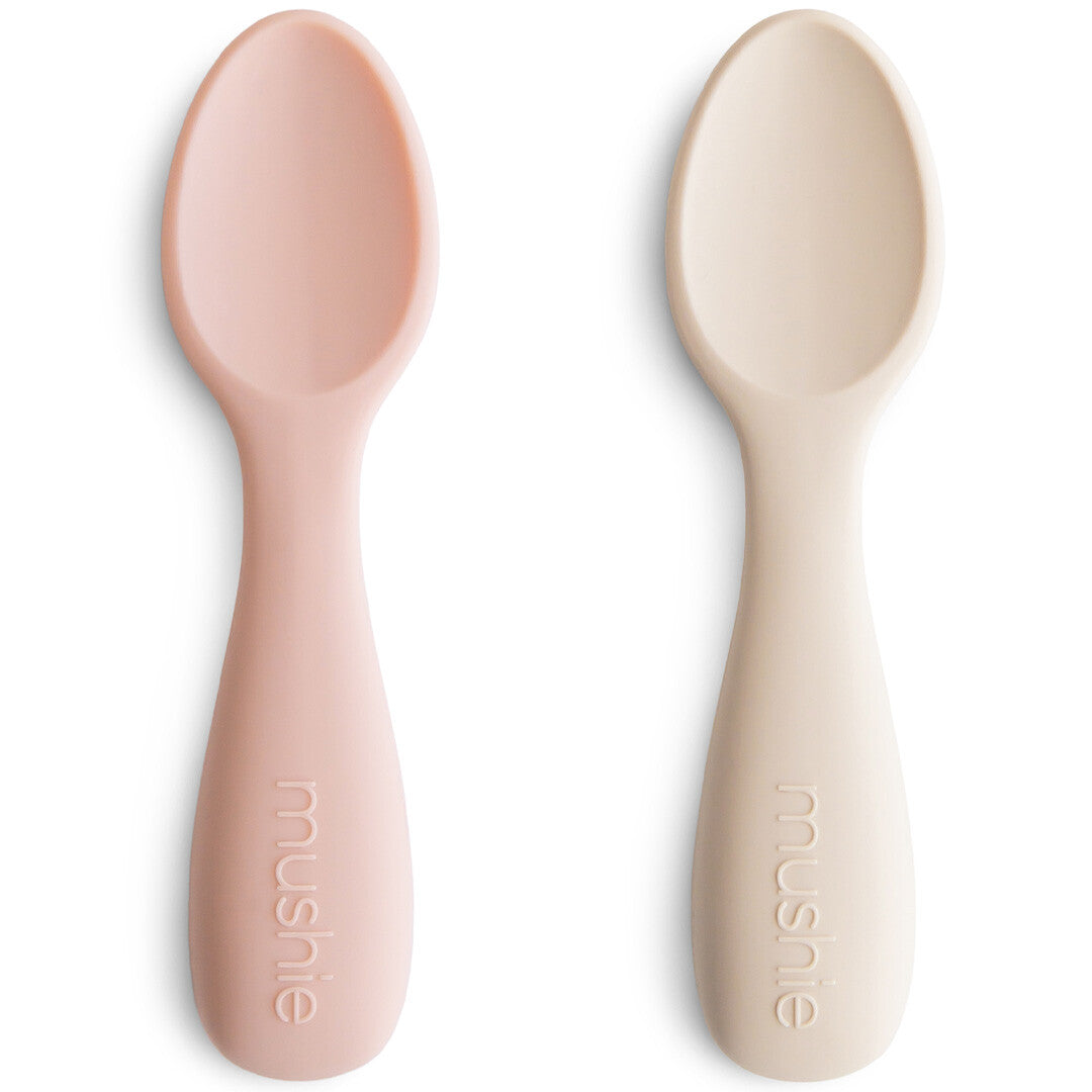 Twee mushie siliconen kinderlepels in roze en beige op een witte achtergrond.