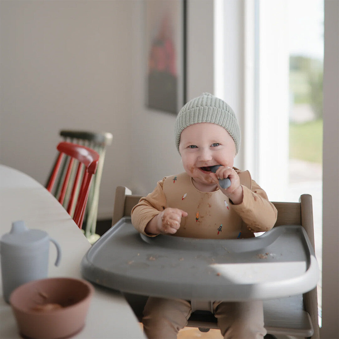 Een lachende baby met een mutsje en slabbetje zit in een kinderstoel en eet met een lepel.