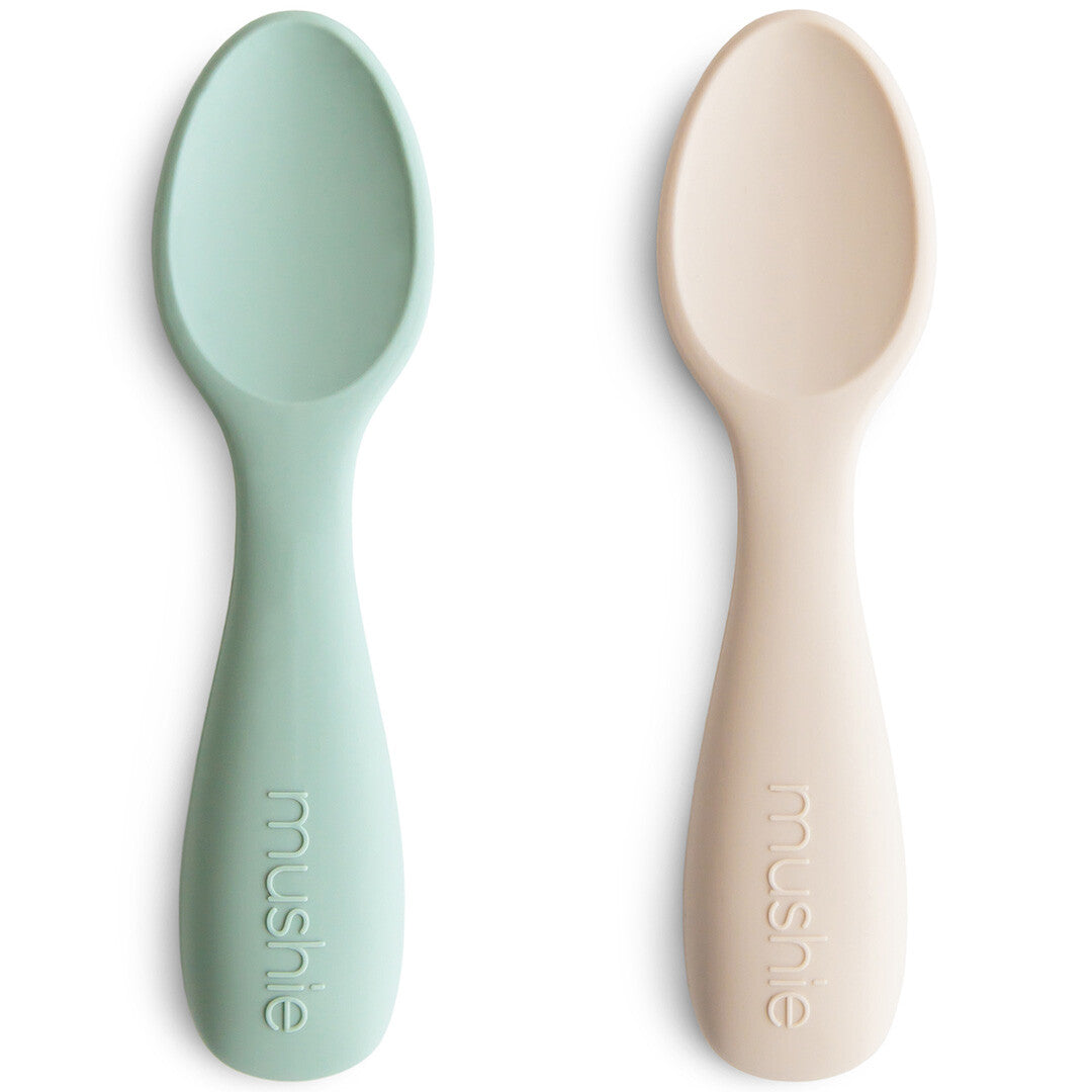 Twee Mushie siliconen baby lepels in lichtblauw en lichtbeige liggen naast elkaar op een witte achtergrond.