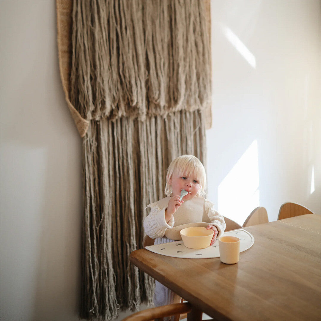 Een peuter in een kinderstoel eet met een Mushie baby lepel uit een gele kom.