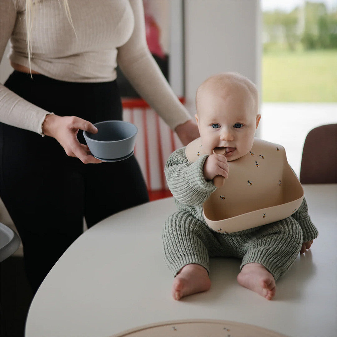 Een baby met een Mushie baby lepel in de mond zit op tafel, met een volwassene die een kom vasthoudt.