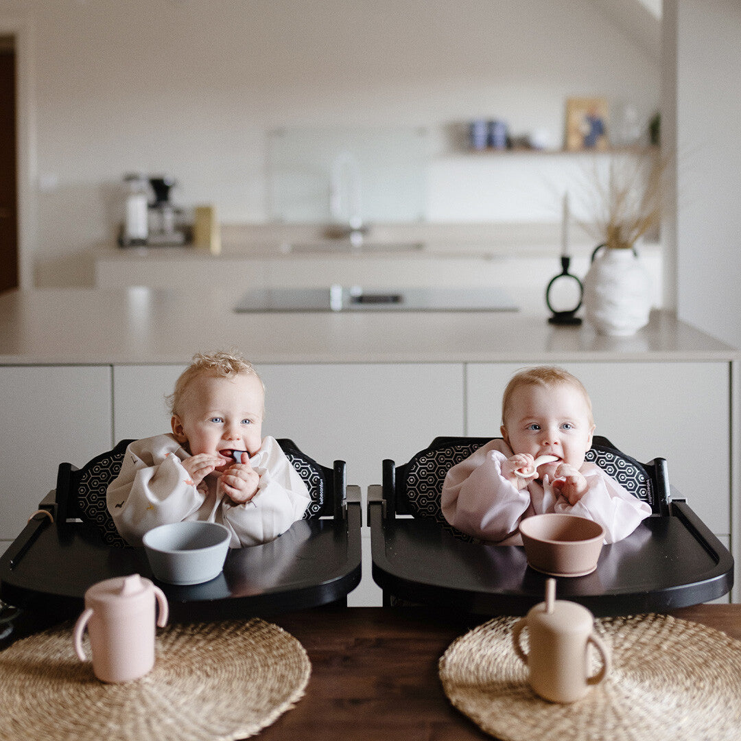 Twee baby's zitten in kinderstoelen aan tafel en eten met Mushie siliconen lepels uit kommen.