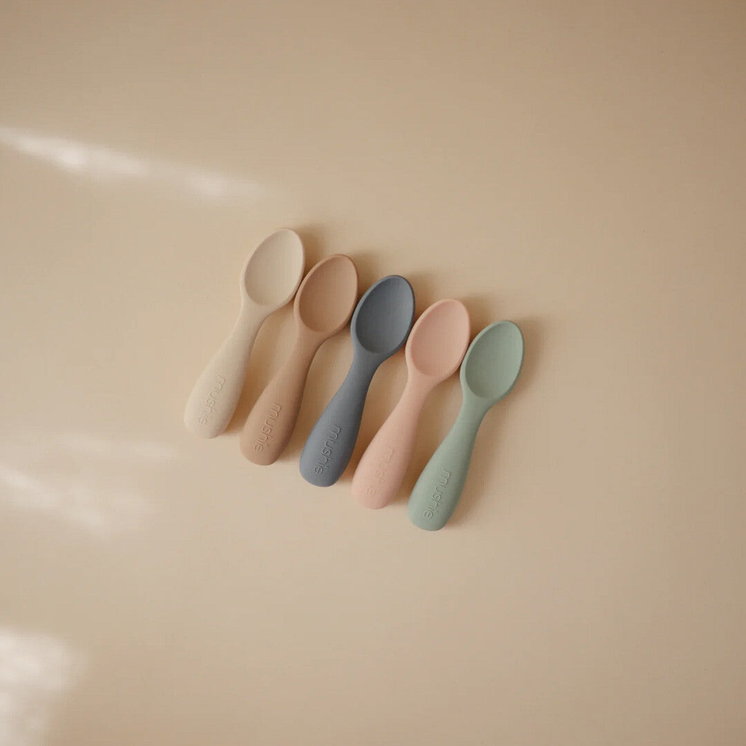 Vijf Mushie siliconen voederlepels in verschillende pastelkleuren liggen op een beige ondergrond.