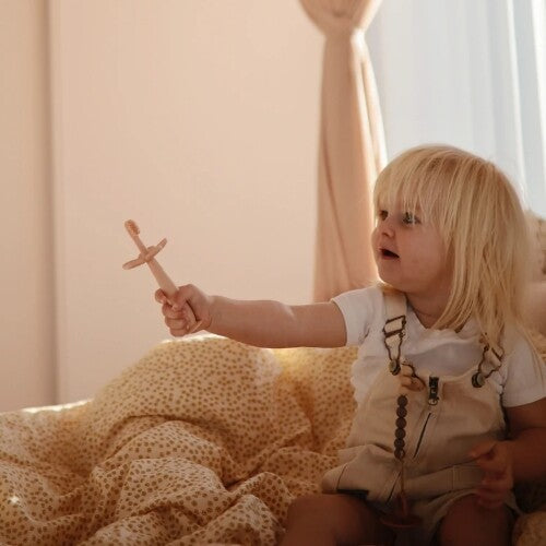 Een kind met blond haar houdt een Mushie siliconen kindertandenborstel vast terwijl het op een bed zit.