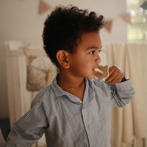 Een peuter met krullend haar gebruikt een beige mushie kindertandenborstel met stervormige bescherming.