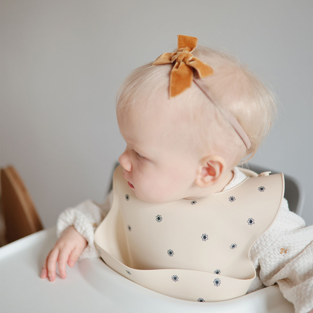 Een baby draagt een beige siliconen slabbetje met zwart madeliefjespatroon in een kinderstoel.